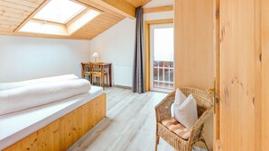 A room with a wooden ceiling, a bed, a wicker chair, and a small table with a lamp.