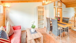 A cozy room with a wooden ceiling, a red sofa with American flag pillows, a wooden dining table with wicker chairs, and a white wall with a clock.