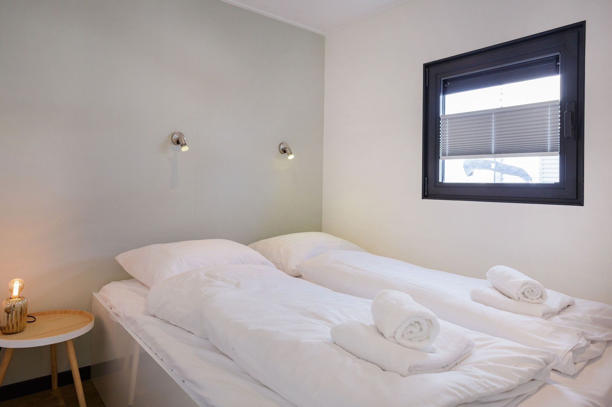 A clean, minimalist hotel room with two single beds, a small bedside table with a lamp, and a window with blinds.