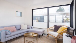 Ein modernes Wohnzimmer mit einem grauen Sofa, einem Korbstuhl, einem kleinen Couchtisch und einem großen Fenster mit Blick ins Freie.
