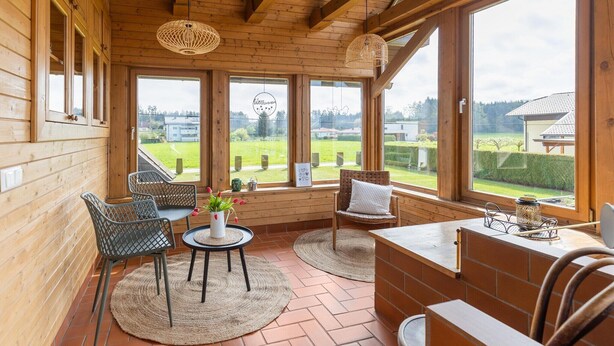 Een houten interieur met grote ramen, een eethoek met stoelen en een tafel, en uitzicht op het omliggende landschap.