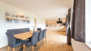 A modern dining area with a wooden table and grey upholstered chairs, a painting on the wall, and a kitchen in the background.