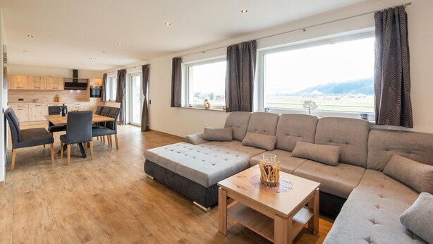 A modern living room with a sectional sofa, a dining table with chairs, and a kitchen in the background.