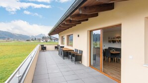 A balcony with a view of a green field and mountains, wooden tables and chairs, and a glass door leading inside.