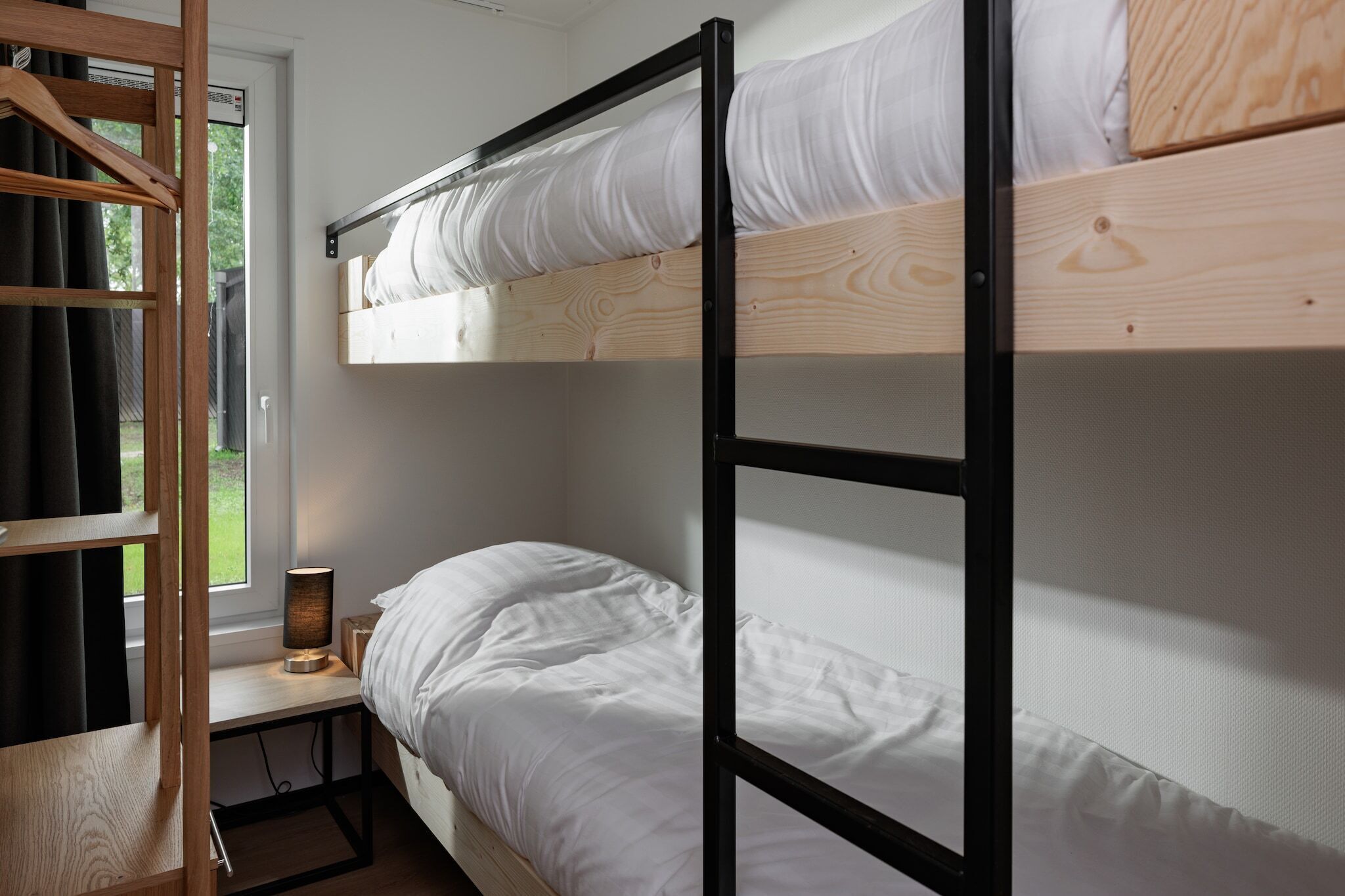A bunk bed with white bedding, a bedside table with a lamp, and a window with curtains.