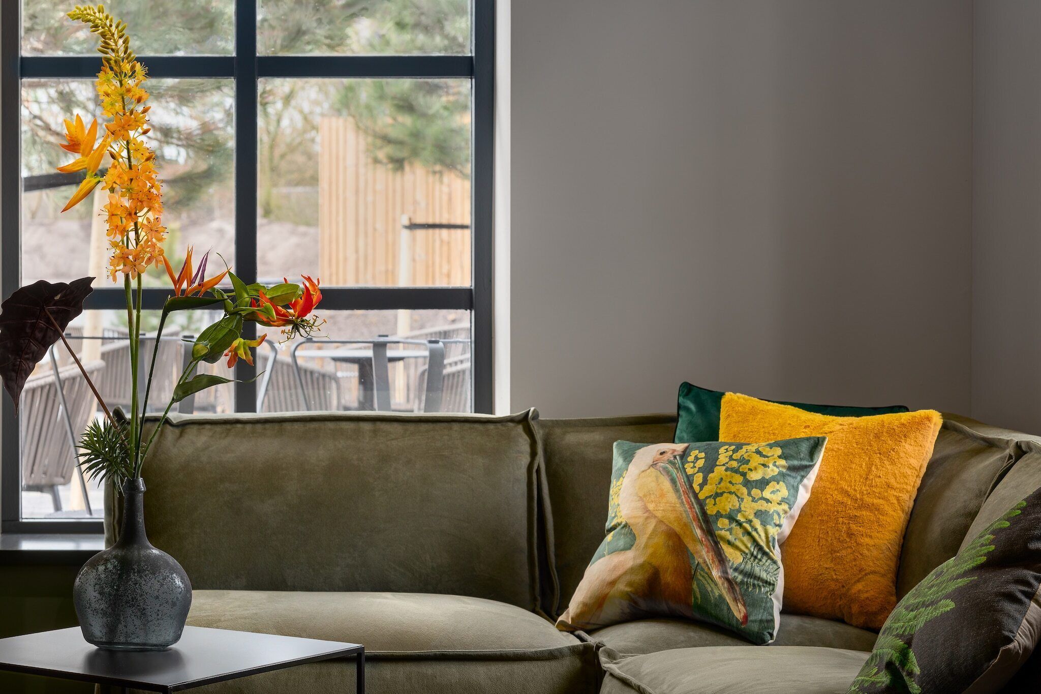 Una sala de estar con un sofá verde, un jarrón con flores y una ventana que da a una valla.