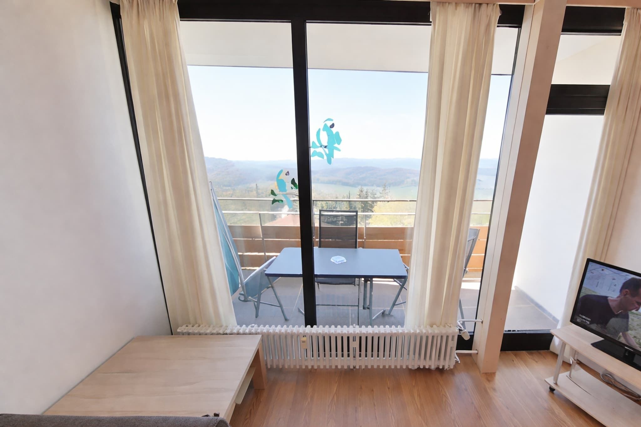 Ein Zimmer mit großem Fenster, Heizkörper, Tisch und Fernseher.
