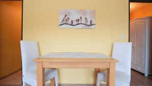 Un comedor con una mesa de madera y sillas blancas, un cuadro de pájaros en la pared y una puerta con un armario al fondo.