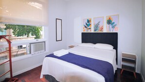 Ett hotellrum med en säng, ett fönster med persienner, ett element och konstverk på väggen.