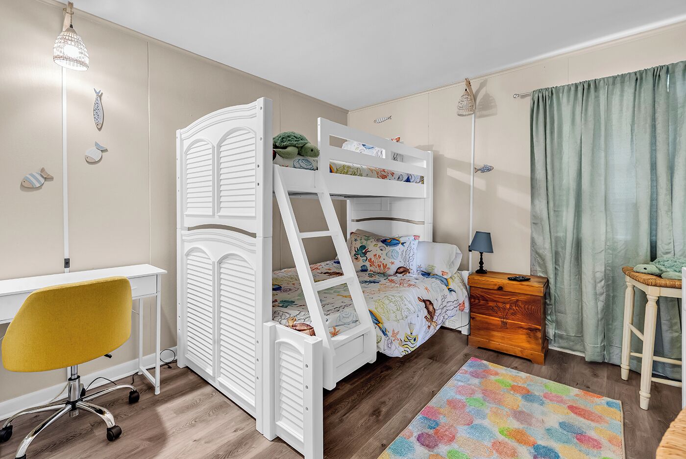 Een kinderbed met stapelbed, een ladder, een bureau, een gele stoel, een houten nachtkastje, een kleurrijk vloerkleed en een gordijn.