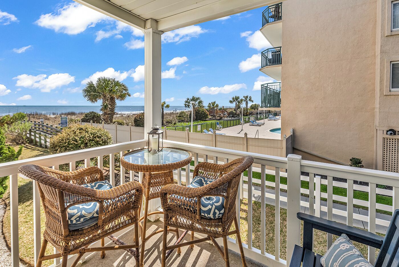 A balcony with wicker chairs and a table, overlooking a beach and ocean.