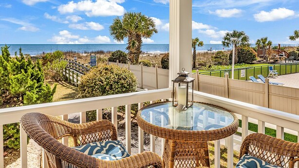 Un balcon avec des fauteuils en osier, une table en verre et une vue sur la plage et l’océan.