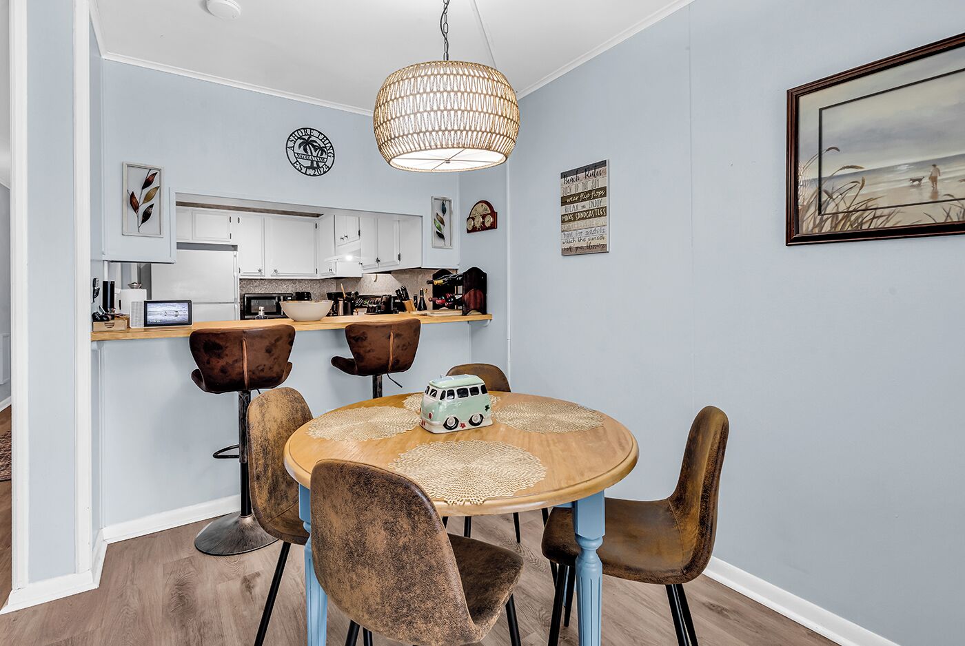 A dining area with a round table, four chairs, a hanging light fixture, and a kitchen in the background.