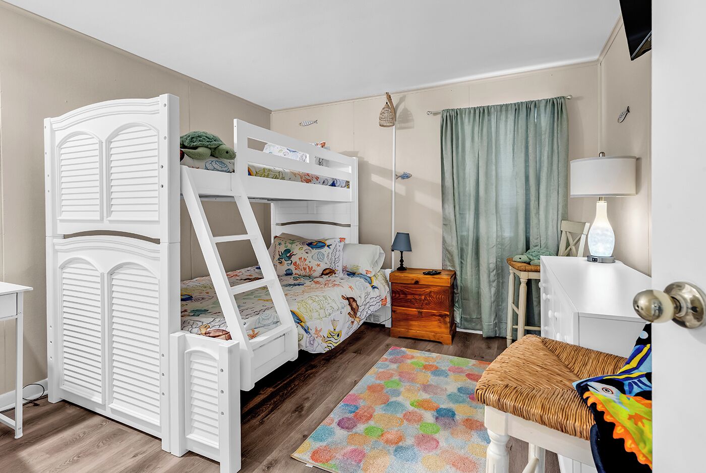 Ein Kinderstockbett mit einer Leiter, ein Nachttisch mit Lampe, eine kleine Holzkiste, ein farbenfroher Teppich und ein Schreibtisch ebenfalls mit Lampe.