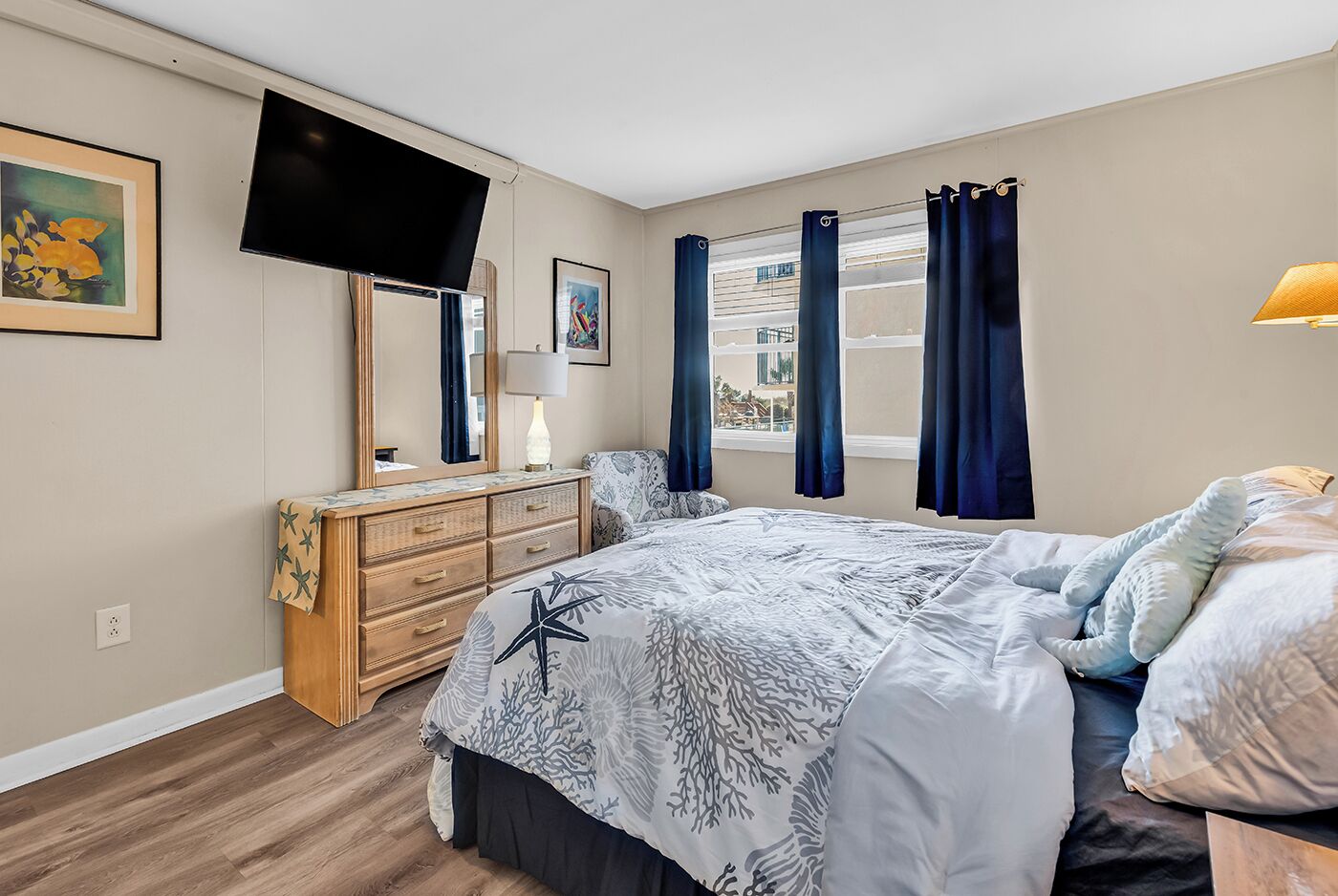 Ein Schlafzimmer mit Bett, Kommode, Fernseher, Fenster mit Vorhängen und einem Bild an der Wand.