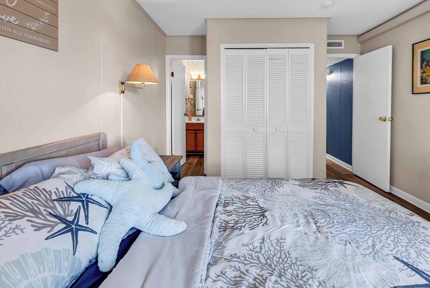 A bedroom with a bed, a nightstand, a lamp, a closet, and a bathroom visible in the background.