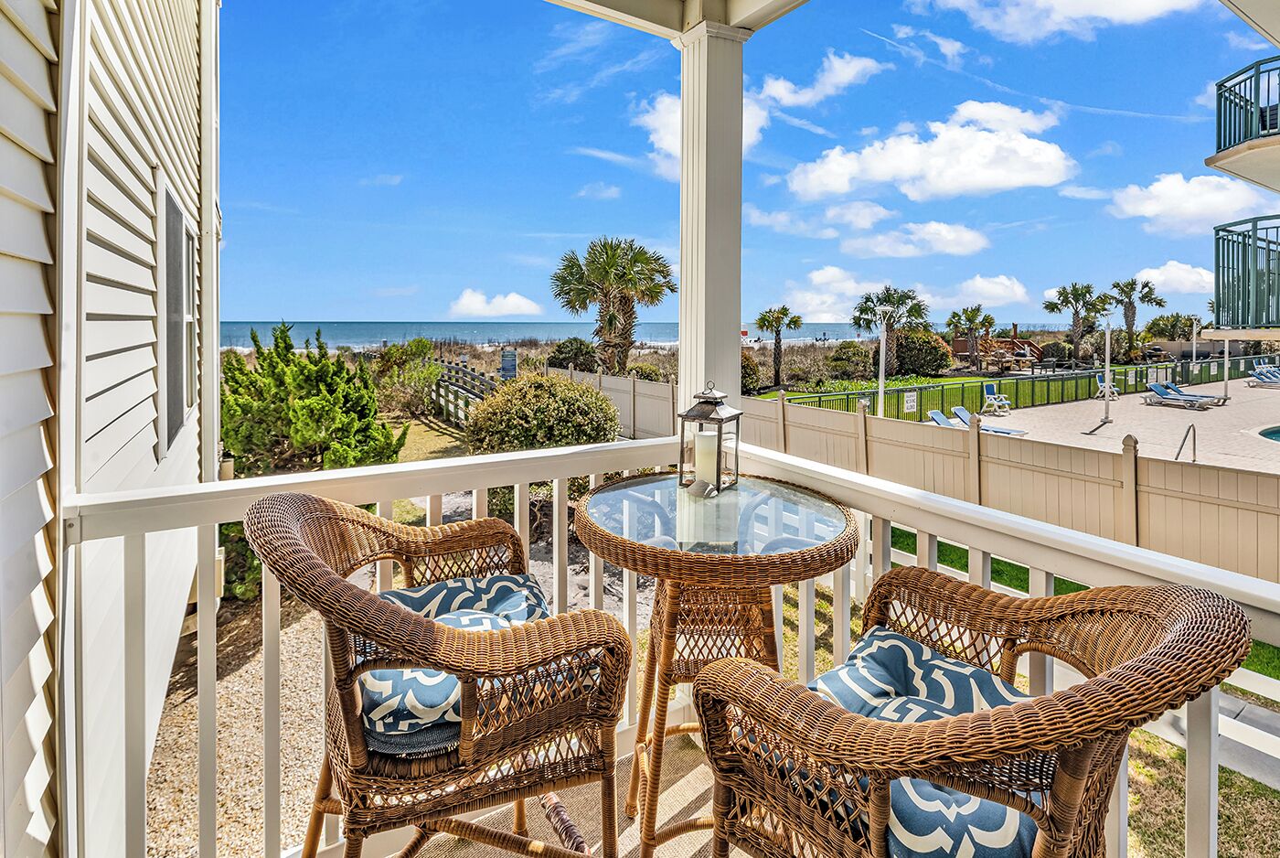 A balcony with wicker chairs and a glass table, overlooking a beach and ocean.
