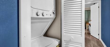 A laundry area with stacked washers and dryers, a door with a white handle, and a hallway leading to a room with a wooden floor and a green curtain.