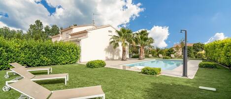 Un patio trasero con piscina, sillas de descanso, palmeras y un edificio blanco.