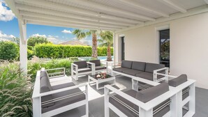 A covered outdoor seating area with white furniture and a view of a swimming pool and greenery. - Villa w/ Palm Trees & Pool (Saint geniès de Fontedit)