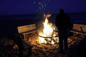 En person står ved et lejrbål på en sandstrand med træbænke.