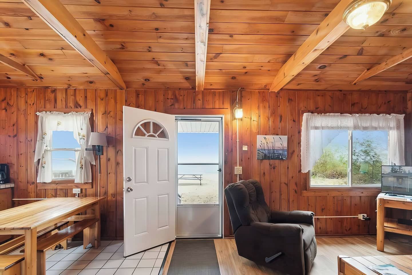 A wooden cabin interior with a dining table, a sofa, a television, and a door leading to an outdoor area.