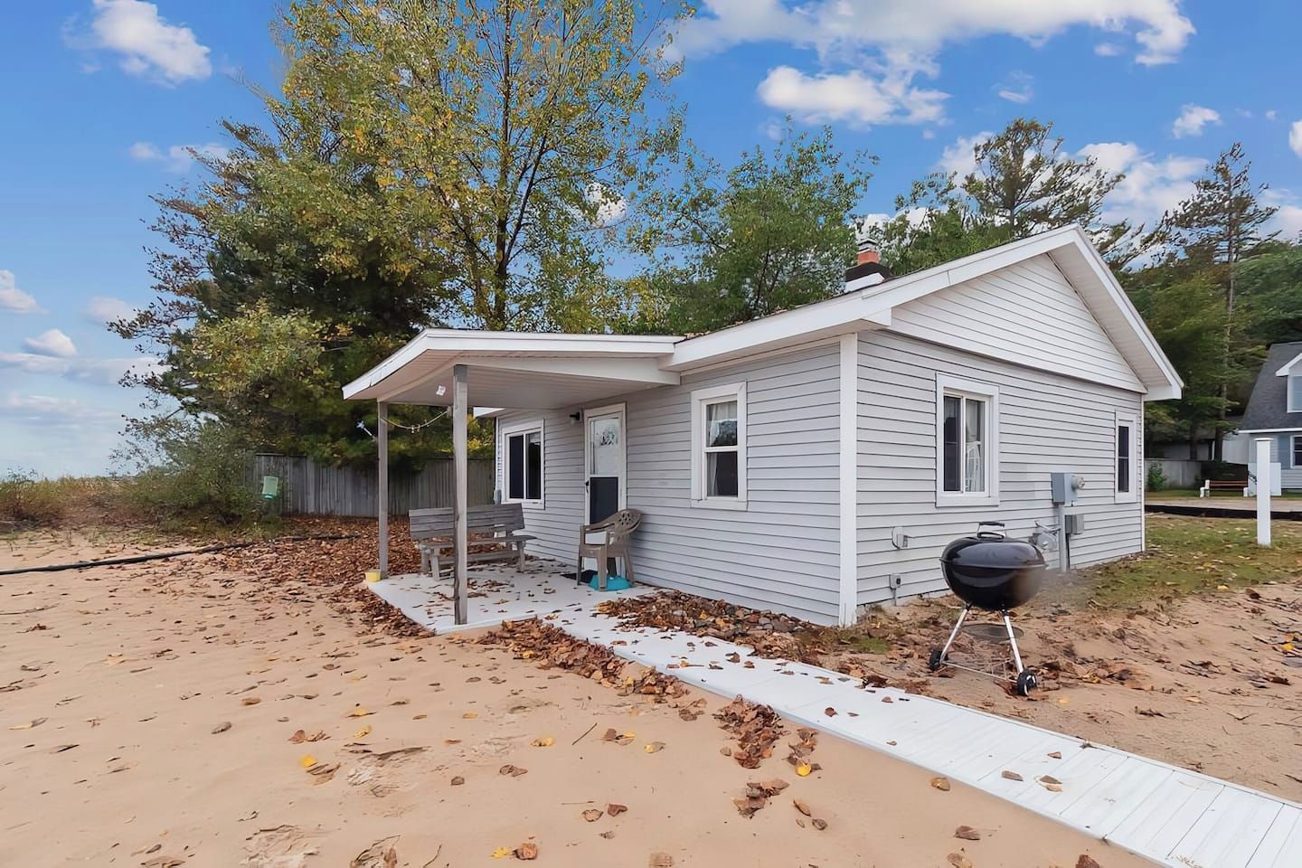 A small, single-story house with a covered porch, a barbecue grill, and a sandy beach area.