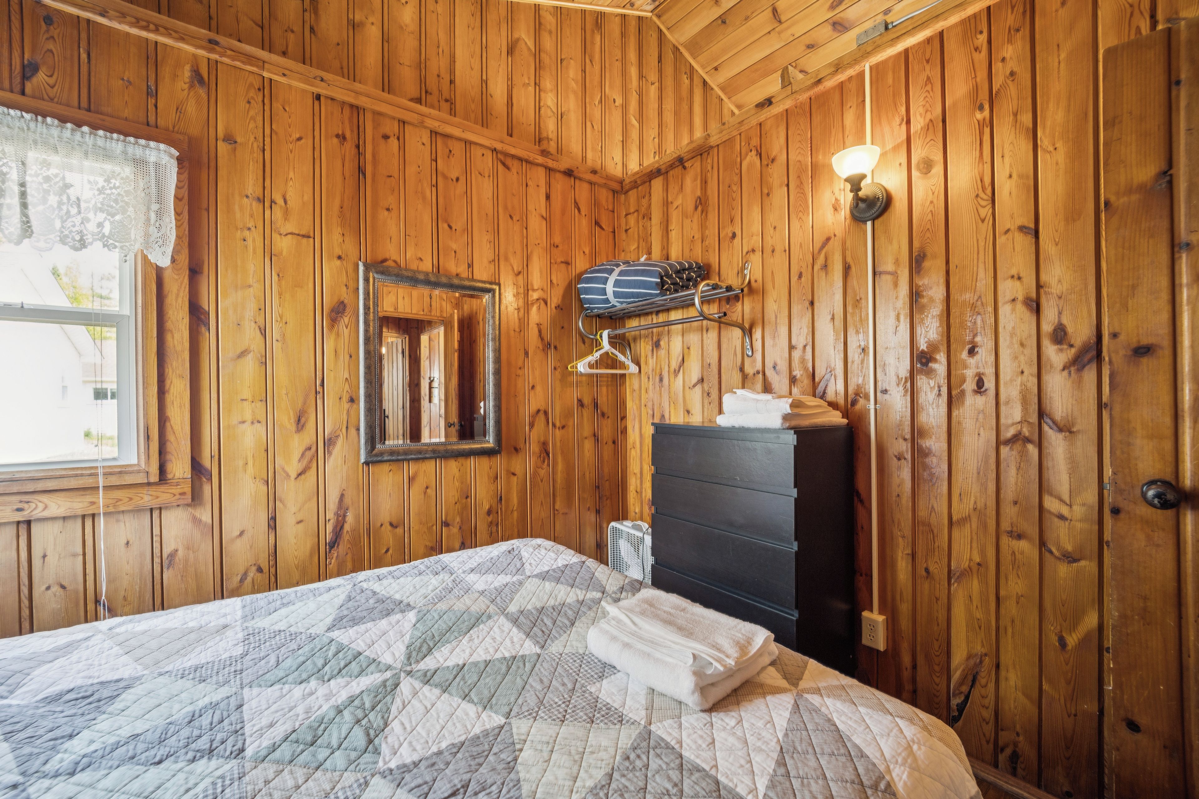 Une pièce aux murs lambrés, comprenant un lit, une commode, un miroir et une lampe fixée au mur.