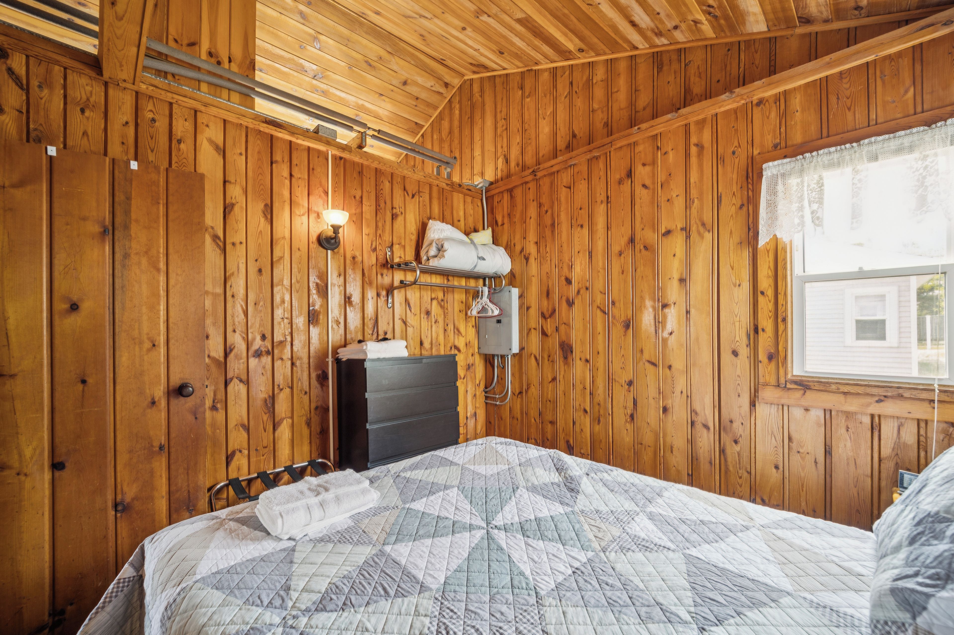 Habitación con paneles de madera, una cama, un aparador, un calentador en la pared y una ventana con vistas a una casa.