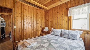 Et rum med træpaneler, en seng, et natbord, et vindue med blonde gardiner og en dør til et skab.