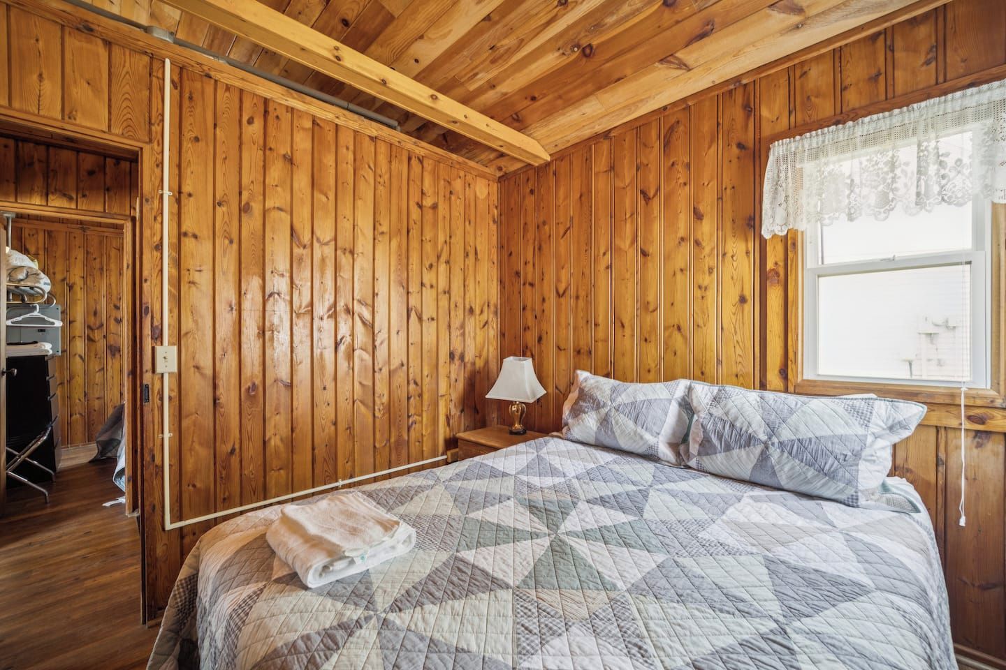 Habitación con paneles de madera, una cama, una mesita de noche, una lámpara y un ventanal con cortinas de encaje.
