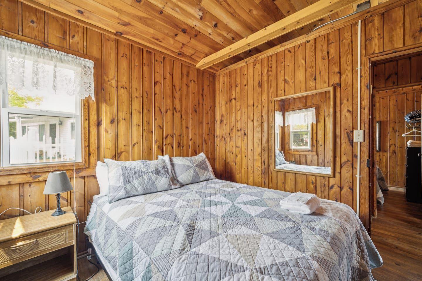 A wooden-paneled bedroom with a bed, a bedside table, a lamp, and a window with a view.