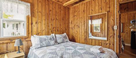 A wooden-paneled bedroom with a bed, a bedside table, a lamp, and a window with a view.