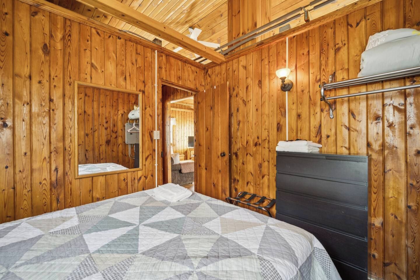 Una habitación con paneles de madera, una cama, un aparador, un espejo y una lámpara fijada a la pared.