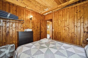 Une pièce aux murs lambrissés, contenant un lit, une commode, un ventilateur et un luminaire.