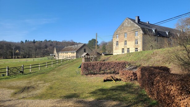 A large, historic building with a prominent chimney, situated on a grassy hill with a wooden fence in the foreground.