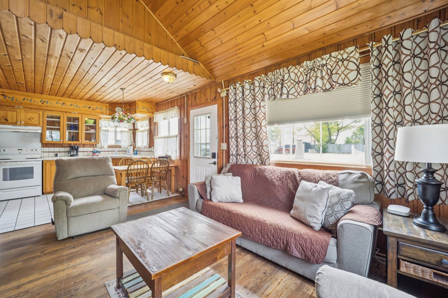 L'interno accogliente di una baita con soffitto in legno, angolo cottura, divano, tavolo da pranzo e tende a motivi decorativi.