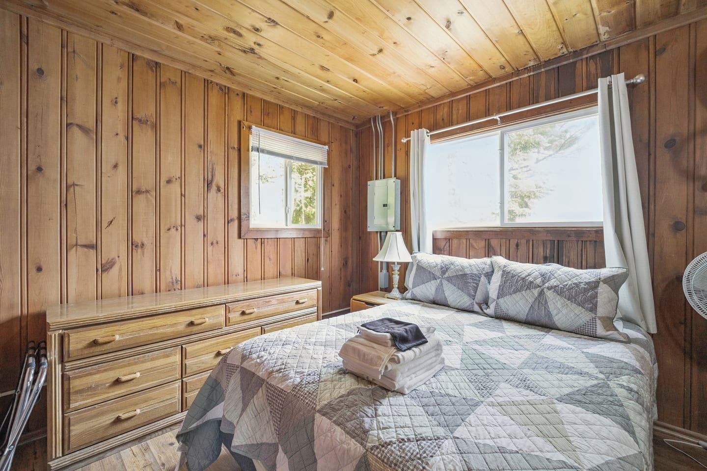 Una habitación con paneles de madera, una cama, un aparador, una ventana y un ventilador.