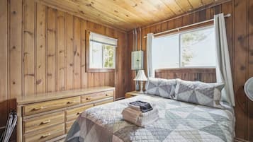 Una habitación con paneles de madera, una cama, un aparador, una ventana y un ventilador.