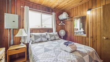 A wooden-paneled room with a bed, a bedside table with a lamp, a wall-mounted heater, a fan, and a mirror.