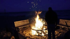 En person står vid ett lägerbål på en sandstrand, och i närheten finns träbänkar.