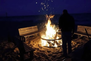 Iemand staat bij een kampvuur op een zanderig strand, met houten banken.