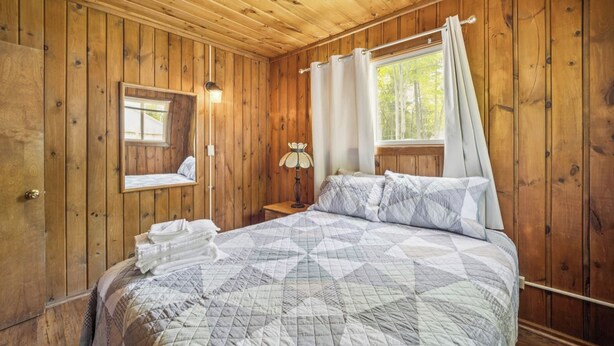 Ett sovrum i en stuga med träpanel, inrett med en säng, ett nattduksbord, en lampa och ett fönster med gardiner.