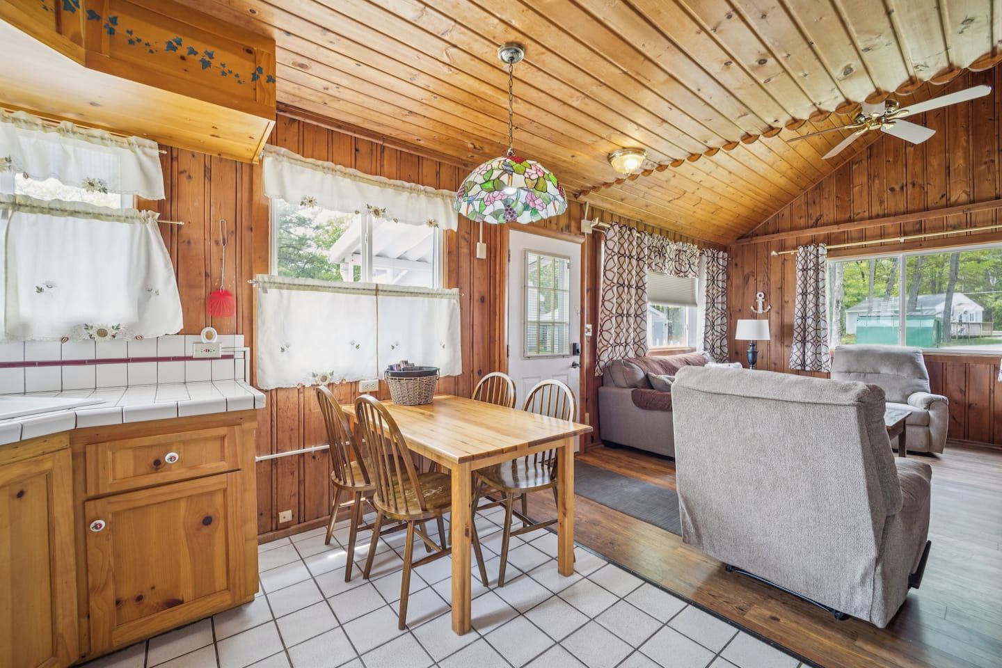 A wooden cabin interior with a dining area, a sofa, and a window overlooking greenery.