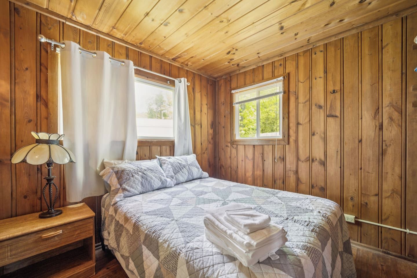 Una habitación con paneles de madera, una cama, una mesita de noche, una lámpara y una ventana con cortinas.