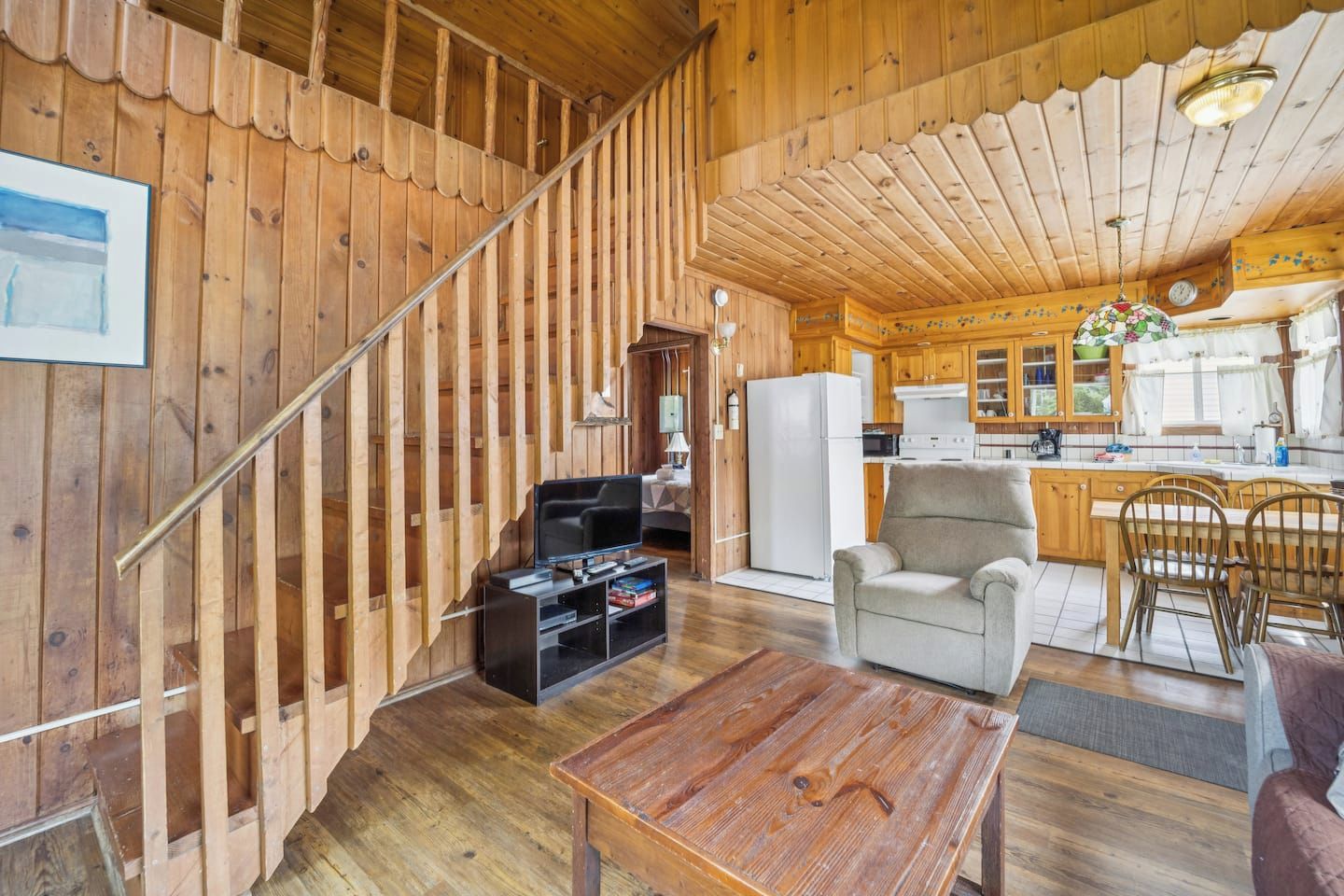 Un intérieur en bois avec un escalier, un espace de vie comprenant un canapé et un téléviseur, une cuisine avec des meubles en bois et un coin repas avec des chaises.