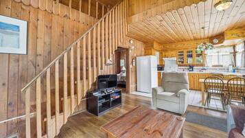 Un intérieur en bois avec un escalier, un espace de vie comprenant un canapé et un téléviseur, une cuisine avec des meubles en bois et un coin repas avec des chaises.
