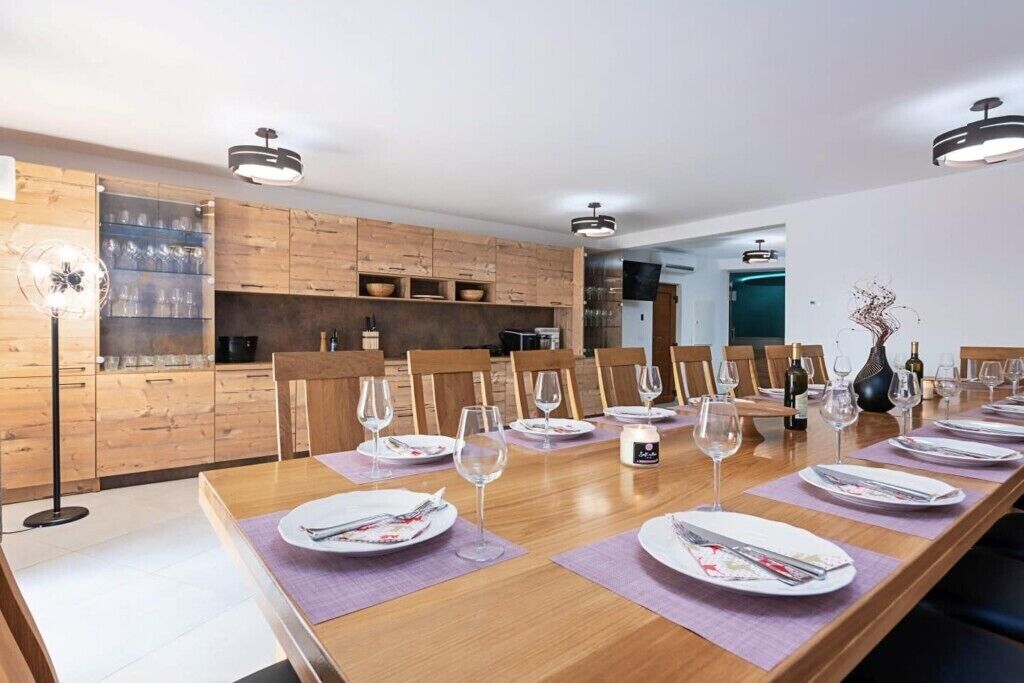 A modern dining area with a long wooden table set for a meal, surrounded by wooden cabinets and a kitchen in the background.