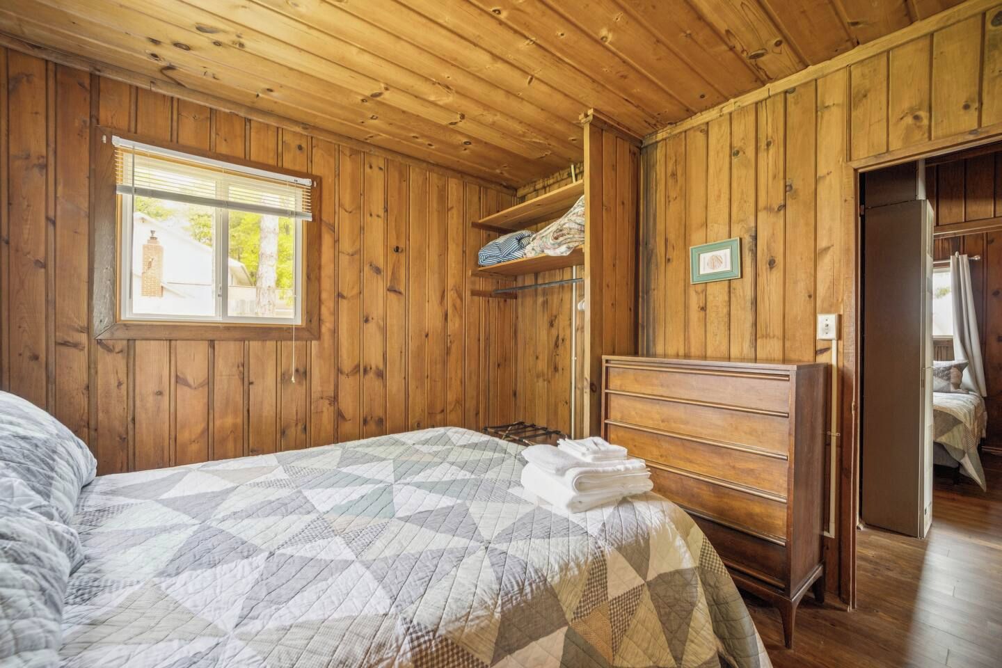 Ein Zimmer mit Holzwänden, einem Bett, einem Schminktisch und einem Fenster mit Jalousien.