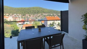 A balcony with a table and chairs, overlooking a town with red-roofed buildings and a hill in the distance.